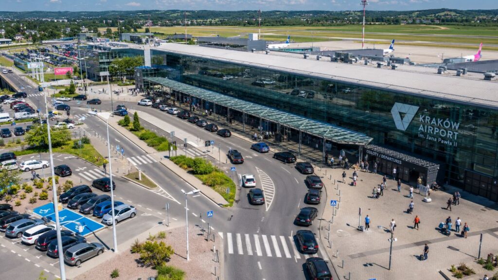 Widok na terminal lotniska Kraków-Balice z lotu ptaka w słoneczny dzień, z widocznym ruchem samochodowym i pasażerami zmierzającymi do wejścia